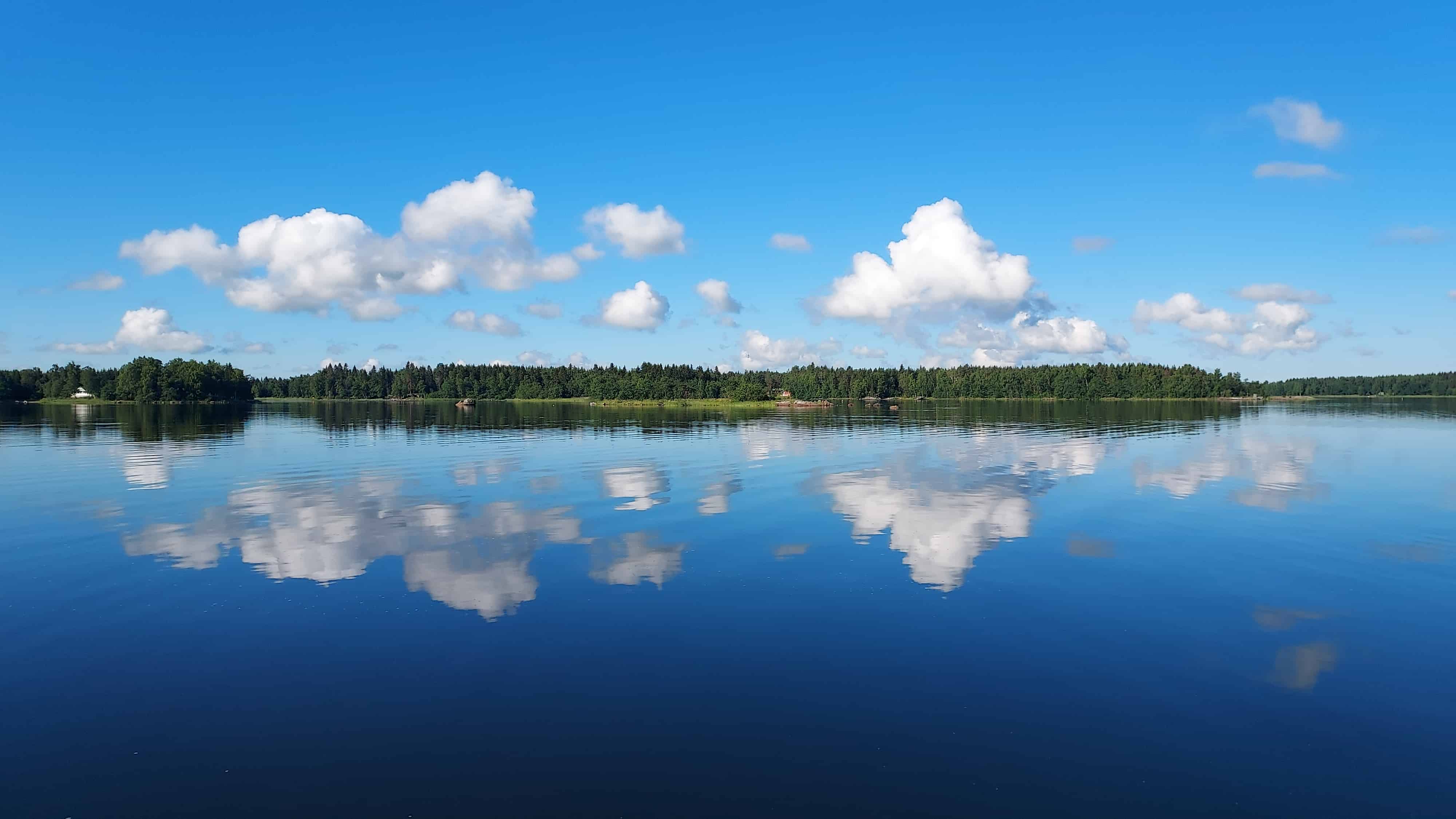 Merimaisema kuvattuna Isokarin saarelta. Meri on tyyni ja horisontissa näkyy metsää. Kirkkaan sinisellä taivaalla on muutamia pilviä, joista heijastuvat kuvat meren pintaan.