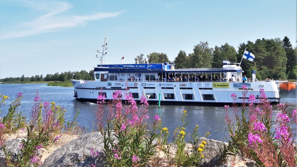 En somrig vy över skärgårdskryssaren M/S Jenny som närmar sig stranden. I förgrunden syns strandstenar bland vilka blommor i olika färger växer. Vita Jenny är full av passagerare och i aktern vajar Finlands flagga. Bakom fartyget syns motsatta stranden med sina träd.