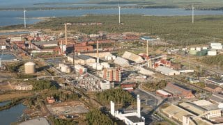 Vy över Karleby storindustriområde med sina fabriksbyggnader sett snett uppifrån. I bakgrunden syns skog med vindkraftverk och längre bort havet.