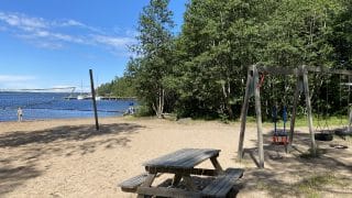 Långö strand i Öja. I bildens vänstra kant syns en strandvolleybollplan och bakom den havet och en badare som vadar i strandvattnet. I mitten, i bildens förgrund, finns ett träbord med bänkar och höger om det trägungor. Det finns många träd i bildens bakgrund.