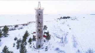 Vintrig bild som tagits snett uppifrån av Harrbåda fyr. Bakom fyren syns små byggnader och längre bort i bakgrunden finns det snötäckta havet.