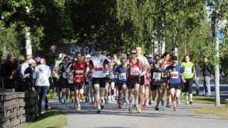 En stor grupp löpare löper i skuggan av träd mot kameran. Löparna har nummerlappar på sina skjortor. Bland löparna finns män, kvinnor och barn. I vänstra kanten finns ett staket bakom vilket det finns människor som ser på löparna.