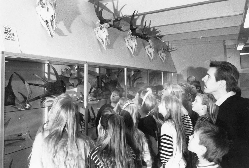 A teacher and students are looking at elk cast antlers on top shelf of a glass cabinet.