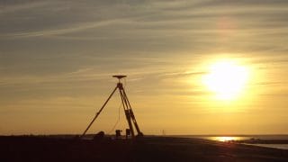 Stödpunktsmätning på stranden. Foto tagen i kvällsskymningen av en GPS-navigator på en öppen strand. I bakgrunden skimrar havet och på himlen sjunker solen bakom en molnslöja.