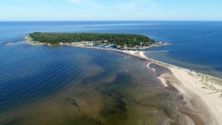 På fotot taget ur fågelperspektiv syns Ohtakari och dess båthamn omgivna av havet. I förgrunden syns en liten bit sandstrand samt den smala vägen som leder till Ohtakari. Bakom Ohtakaris båthamn växer skog.