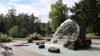 Västraparkens Svallvågsstaty som sprutar vatten i bassängen runt den. Bakom statyn öppnar sig Västraparken med sina träd.