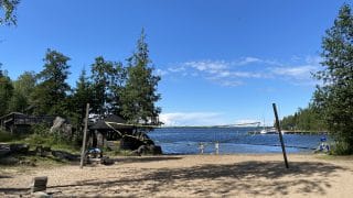 Långö strand i Öja. I bildens förgrund finns sandstrand med en volleybollplan. Bakom planen syns ett blått hav och två badare gående i strandvattnet. Längre bort syns en brygga med båtar. I båda kanterna på bilden syns träd.