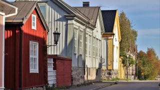 Gatuvy över somrig Neristan. I vänstra kanten gamla trähus, närmast ett rött hus med sadeltak och vita fönsterkarmar. Från husets vägg skjuter ut ett metallskaft med en gammal gatlykta på ändan. Framför husen syns en asfalterad gata.