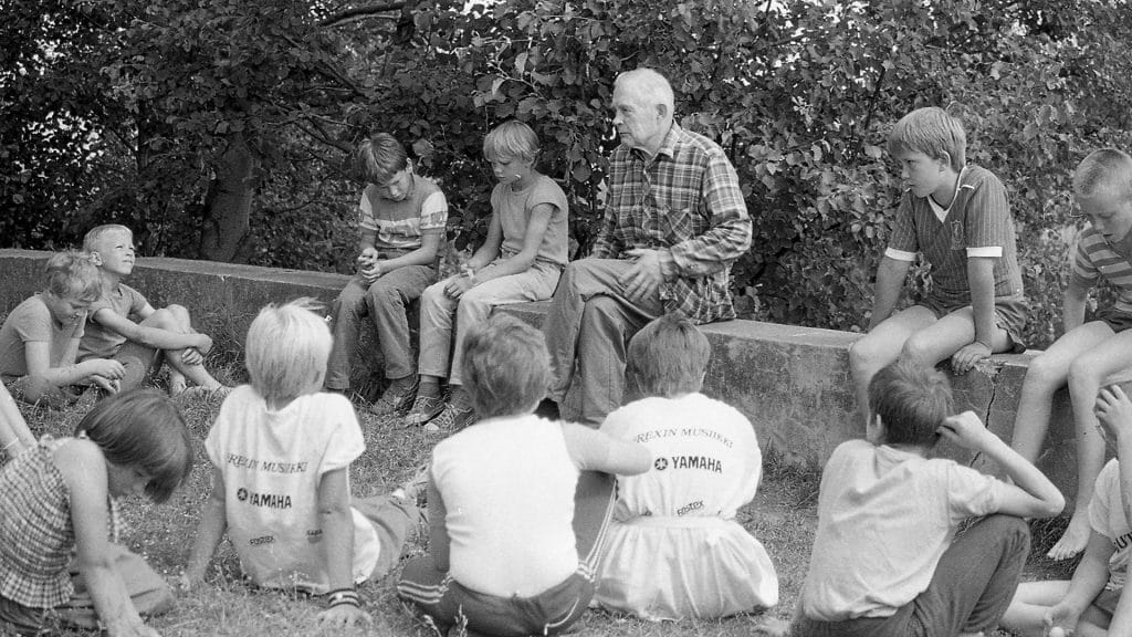 Salkio och fyra barn sitter på en stenmur. På marken framför dem sitter andra barn på gräsmattan.