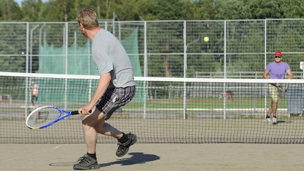 Kaksi miestä pelaamassa tennistä ulkokentällä. Lähempänä, selin kameraan olevalla miehellä on kädessään sininen maila ja yllään ruudulliset shortsit sekä harmaa t-paita. Kauempana, verkon toisella puolella olevalla miehellä on yllään lila t-paita sekä päässään lippalakki ja aurinkolasit. Verkon yläpuolella lentää keltainen tennispallo.