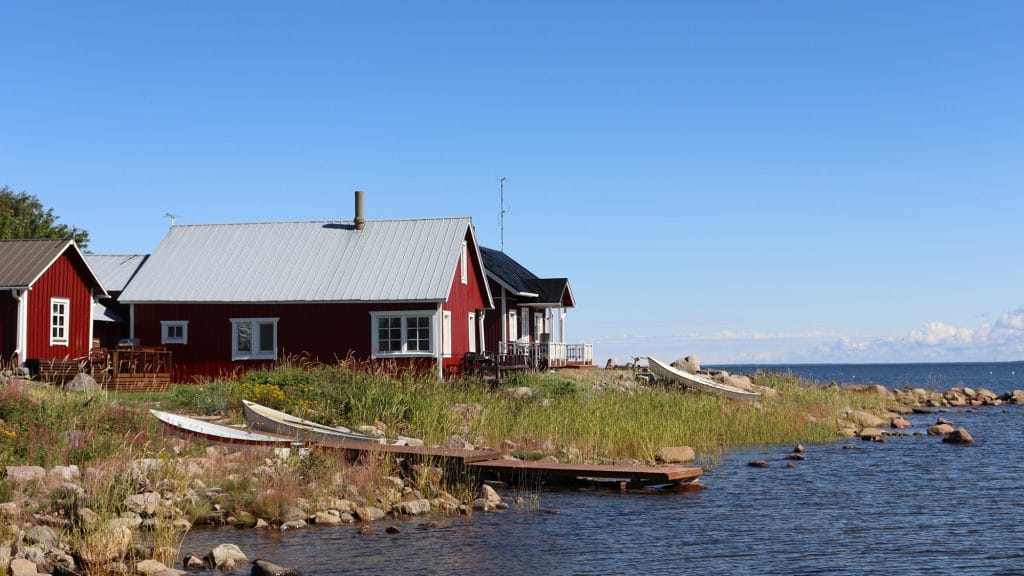 Stenig och gräsbevuxen strandlinje i Ohtakari. I förgrunden en träbrygga som skjuter ut i havet och bredvid två båtar som lyfts upp på stranden. I bakgrunden syns röda byggnader med sadeltak samt en blå, nästan molnfri himmel. I högra kanten syns mörkblått hav.