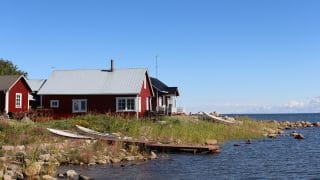Stenig och gräsbevuxen strandlinje i Ohtakari. I förgrunden en träbrygga som skjuter ut i havet och bredvid två båtar som lyfts upp på stranden. I bakgrunden syns röda byggnader med sadeltak samt en blå, nästan molnfri himmel. I högra kanten syns mörkblått hav.