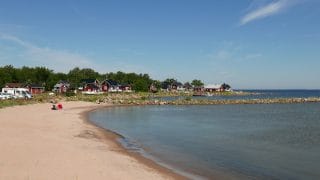 Vy över Vattaja badstrand. I vänstra kanten syns en sandstrand och i högra kanten havet. I bakgrunden finns en vågbrytare av sten som skjuter ut i havet samt stugor med sadeltak och några bilar.
