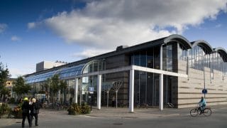 Kokkolan kaupunginkirjasto etuviistosta kuvattuna. Kirjastolla on useista kaarevista elementeistä tehty katto sekä harmaasta kivestä ja suurista lasipinnoista tehdyt seinärakenteet. Kirjaston edestä kulkee Tehtaankatu, jossa liikkuu jalankulkijoita ja pyöräilijöitä. Kadulla on myös runsaasti istutuksia. Kirjaston yläpuolella näkyy sininen taivas, jossa isoja pilviä.