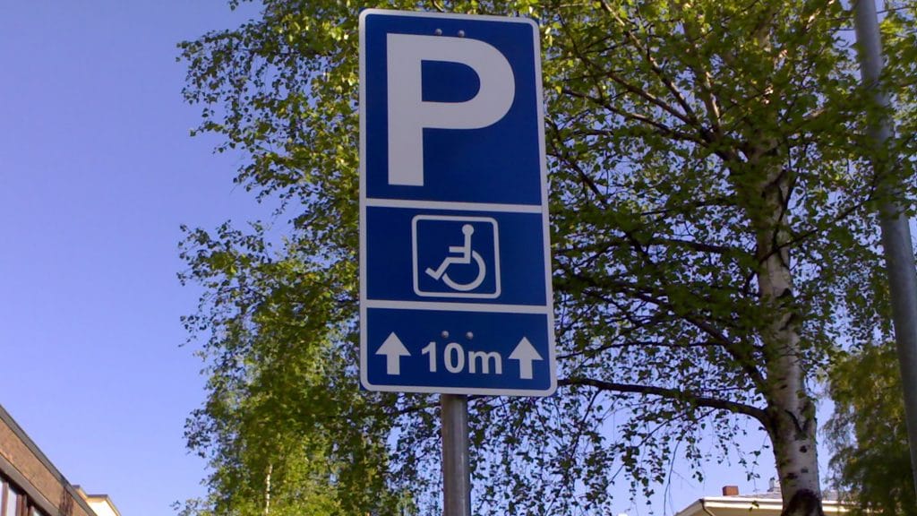 Blåvitt trafikmärke som anger parkeringstillstånd för rörelsehindrade. Bakom märket syns björkar och en blå himmel.
