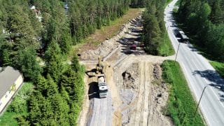 Korkealta ilmasta kuvatty työmaanäkymä, jossa lava-auto on juuri kippaamassa maahan isoa hiekkalastia, jota kasan toisella puolella oleva keltainen kaivinkone on valmiina käsittelemään. Auki kaivetun alueen vierellä kasvaa korkeita puita ja kuvan oikeassa laidassa kulkee asfaltoitu tie. Kuvan vasemmassa laidassa on keltainen, harjakattoinen rakennus.
