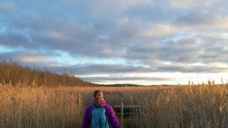 Nainen kulkee luontopolkua kaislikon keskellä reppu selässään.