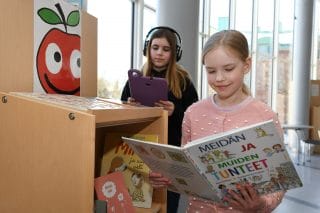 Kaksi nuorta tyttöä seisovat kirjahyllyn luona. Kirjahyllyn päädyssä on ompun kuva. Toinen tytöistä lukee kirjaa ja toinen katsoo kirjaston Hublet-tablettia kuulokkeet korvillaan.