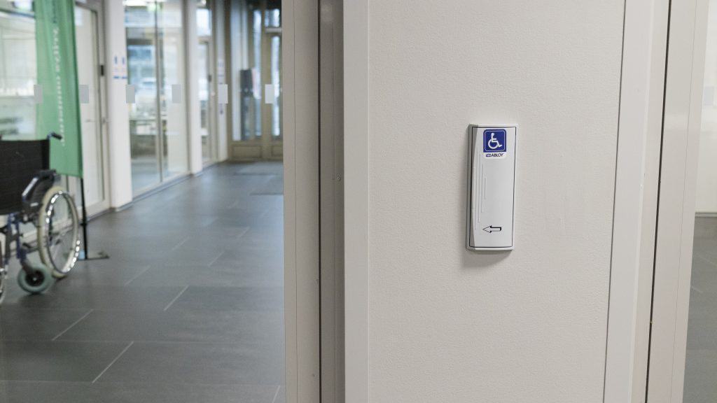 Utsikt från en glasdörr med en automatisk frigöringsknapp på höger sida. I bakgrunden finns en korridor och en rullstol.
