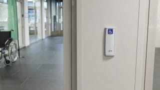 Utsikt från en glasdörr med en automatisk frigöringsknapp på höger sida. I bakgrunden finns en korridor och en rullstol.