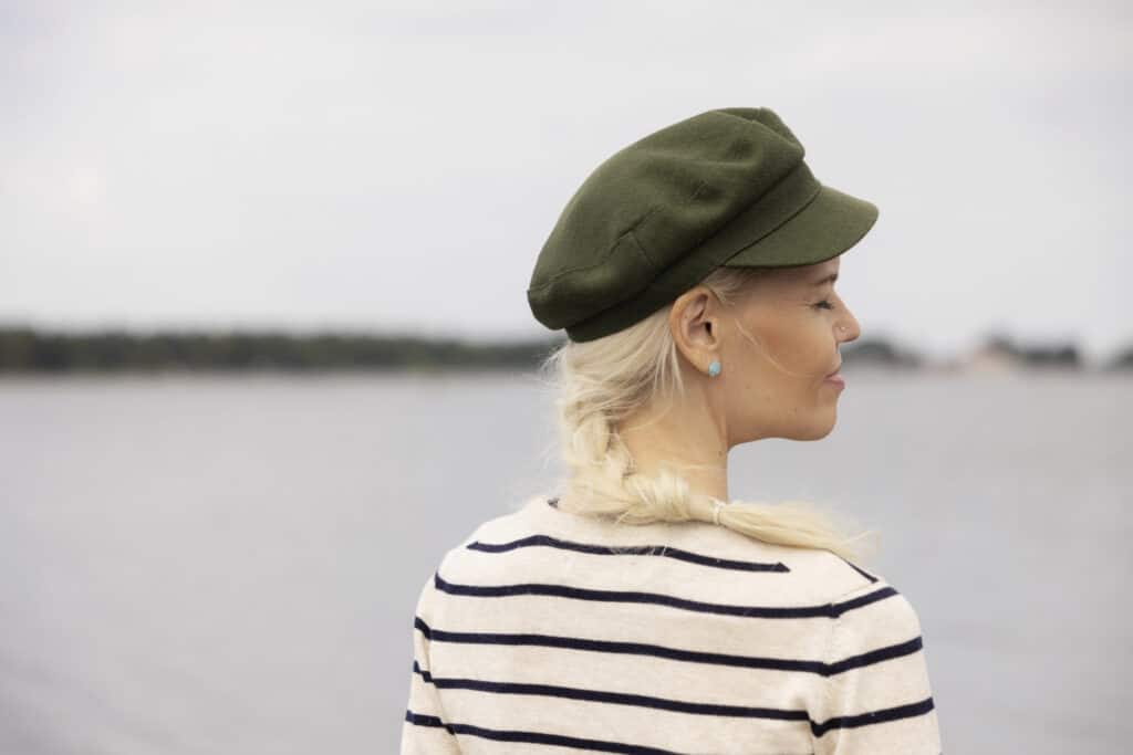 En kvinna klädd i grön keps och svart-vitrandig skjorta står vid havet med ryggen mot fotografen. Kvinnan har långa blonda flätor, hon ler med slutna ögon.