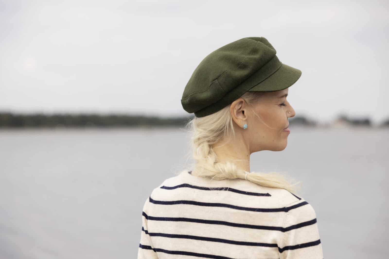 Vihreää lakkiin ja mustavalkoraidalliseen paitaan pukeutunut nainen seisoo meren äärellä. selin kuvaajaan. Naisella on pitkä vaalea letti, hän hymyilee silmät suljettuina.