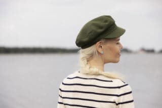 En kvinna klädd i grön keps och svart-vitrandig skjorta står vid havet med ryggen mot fotografen. Kvinnan har långa blonda flätor, hon ler med slutna ögon.