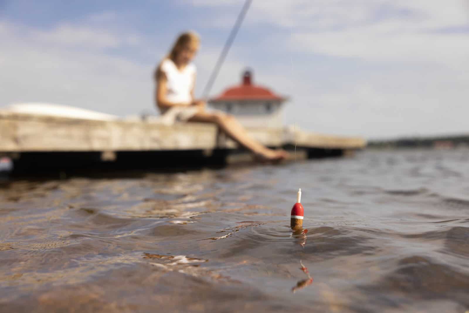 Tyttö istuu laiturilla jalat vedessä ja onkii. Aurinko paistaa. Taustalla näkyy laiturin päähän rakennettu sauna. Ongenkoho keikkuu veden pinnalla.