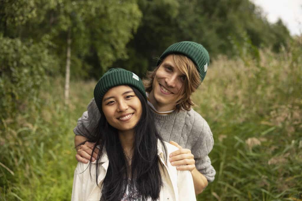 Tumma pitkähiuksinen nuori nainen ja punahiuksinen nuori mies seisovat peräkkäin. Miehen kädet ovat naisen olkapäillä. Molemma hymyilevät ja heillä on päässään vihreät pipot, joissa on teksti Kokkola Karleby. Taustalla näkyy vihreitä kaisloja ja metsää.