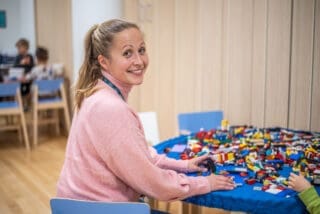 Varhaiskasvatuksen opettaja istuu askartelupöydän edessä, ja katsoo hymyillen kameraan olkansa yli.