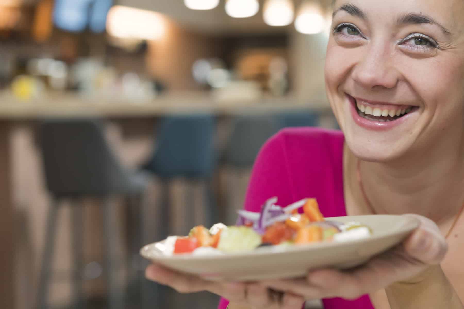 Hymyilevä nainen kohottaa lounaslautastaan kohti katsojaa