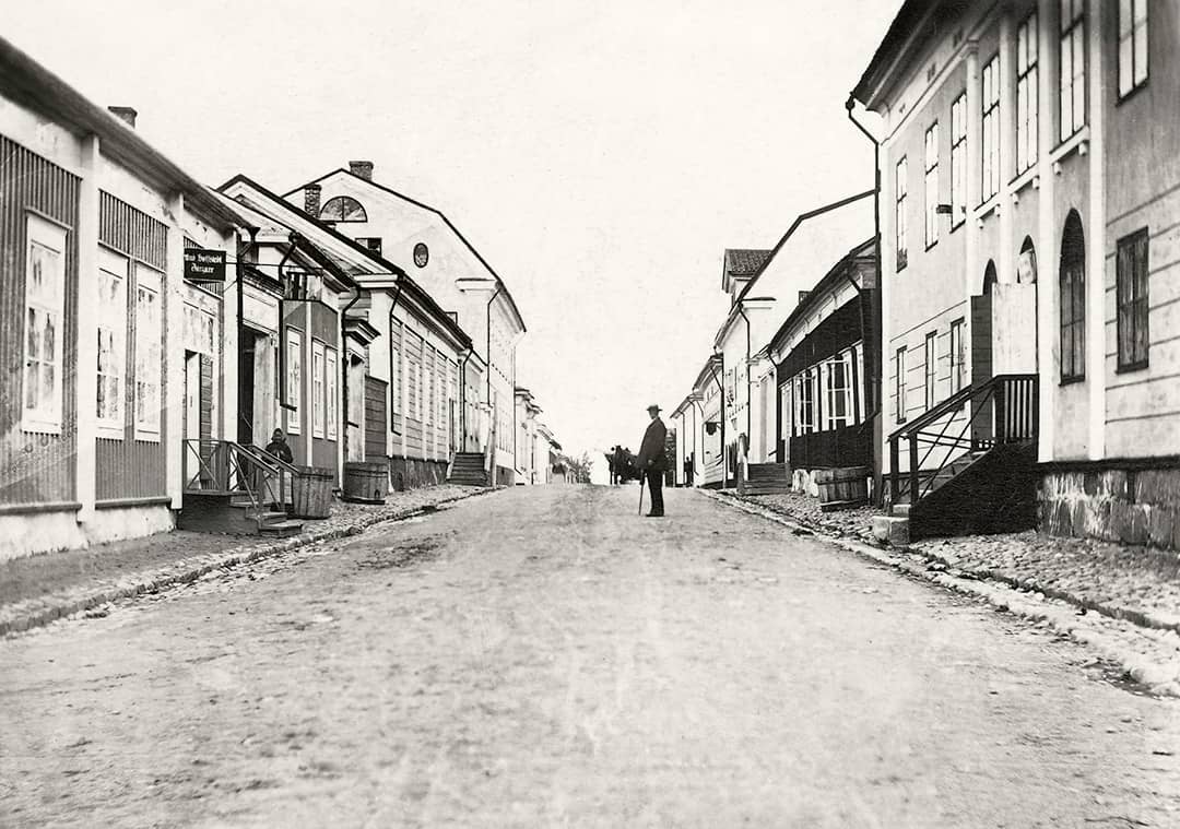 Old back and white photo of Isokatu in Kokkola seen northward from the corner of Tehtaankatu. A man with a walking cane and a hat on his head stands in the middle of the photo.