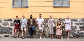 Ryhmä miehiä ja naisia hymyilevät iloisesti Roosin talon seinustan vieressä ja katsovat kameraan.
