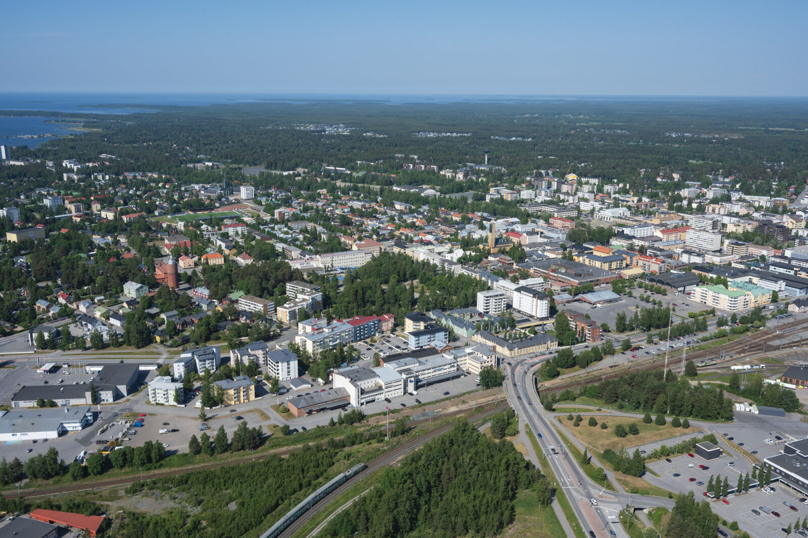 Kuva on otettu korkealta. Etualalla näkyy rautatie ja maantie sekä kevyen liikenteen väyliä, jotka johtavat kaupungin keskustaan. Rakennusten katot näkyvät.