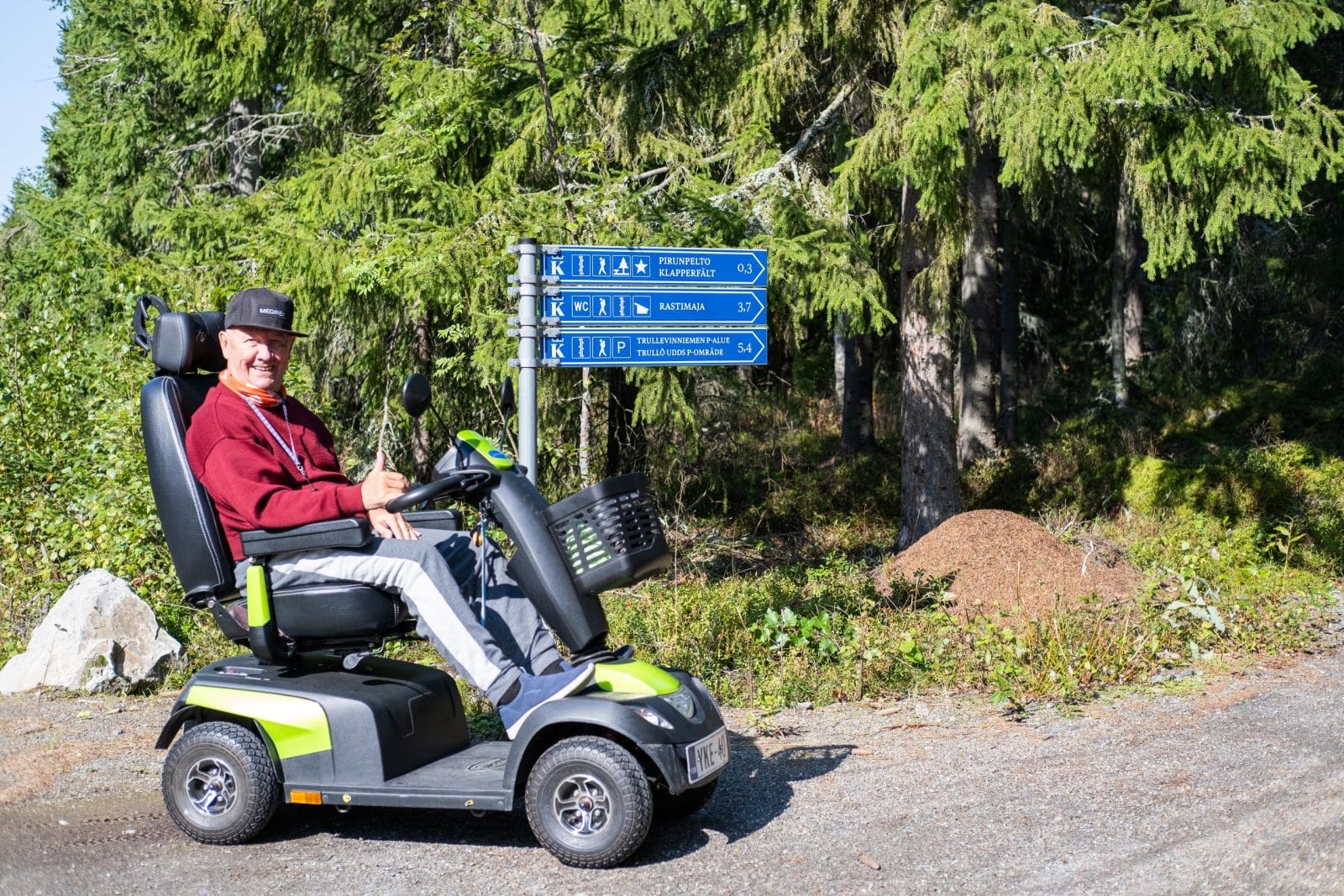 Sähköpyörätuolin käyttäjä hymyilee viittaopasteen vierellä sorastetulla polulla. Viittaopasteen vieressä on muurahaispesä ja takana kuusia.