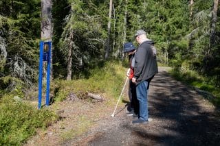 Näköesteinen avustajansa kanssa tutkimassa polun varren luontopolkuopastetta, jossa qr-koodi ja valokuva.