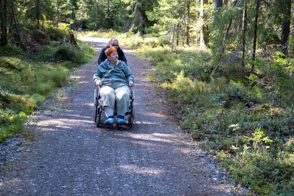 Pyörätuolin käyttäjä avustajineen sorastetulla polulla metsän siimeksessä.
