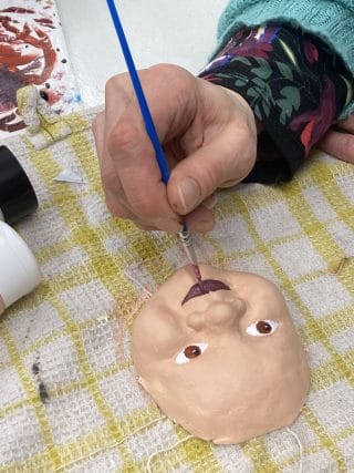 Yksityiskohta kädestä, joka maalaa siveltimellä nuken kasvoja.