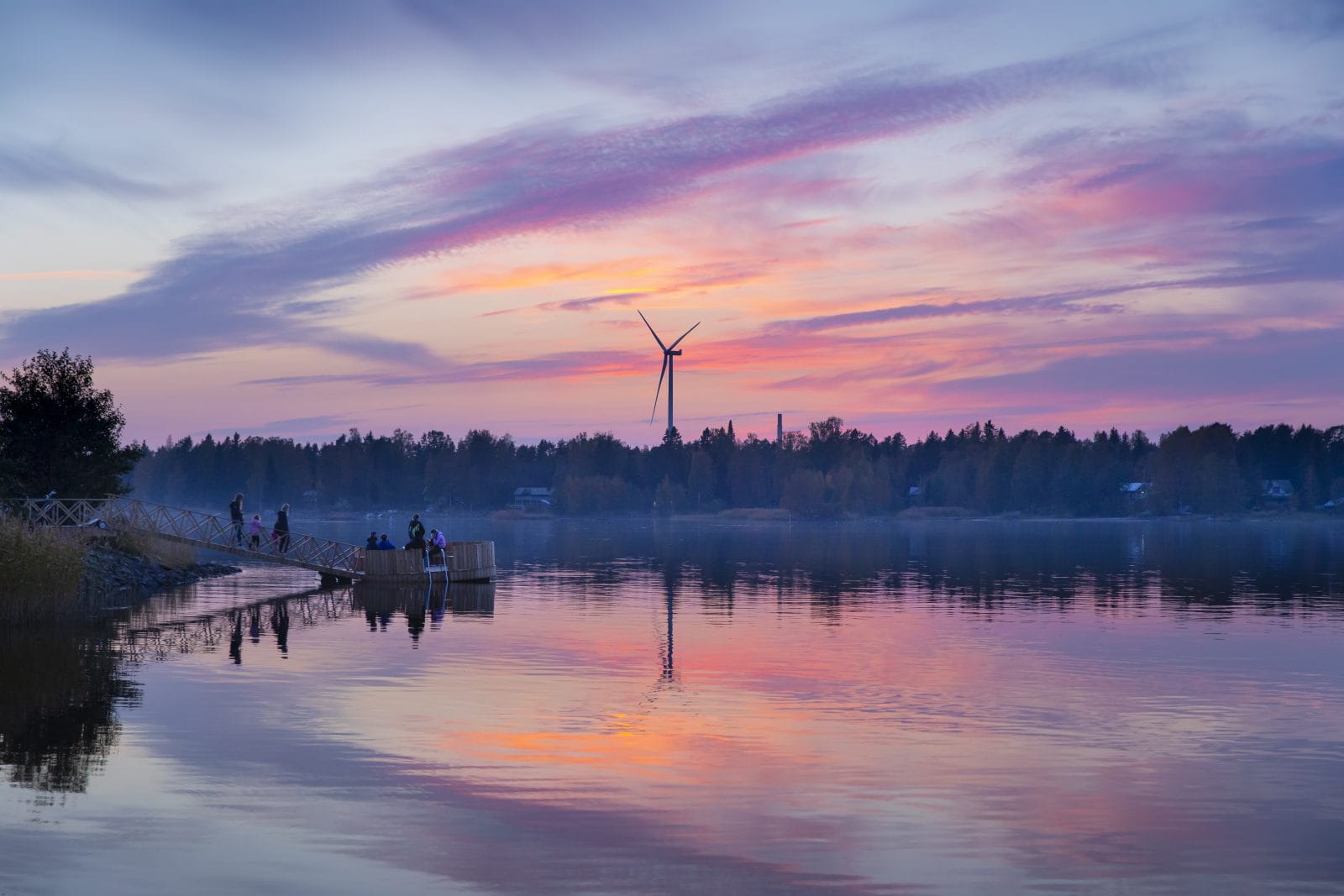 Punasävyiset iltataivaan värit heijastuvat mereen. Takana siintää tumma metsä ja tuulimylly. Rannalla näkyy nuorisoa