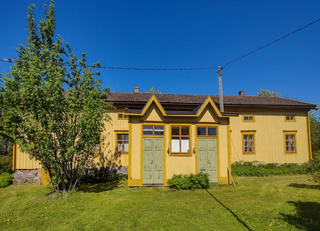 Edestäpäin kuvattuna oleva 1800-luvun puinen keltasävyttäinen Hakalax talo jossa kaksi ovea sisäänkäynneille. Sinen taivas ja talon edessä tasainen nurmikko.
