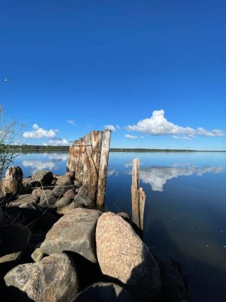 Vanhoja laiturirakenteita rantakivikossa. Meri on tyyni ja pilvet heijastuvat veden pintaan.