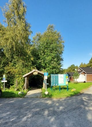 Luontopolulle johtava portti on keskellä kuvaa. Oikealla puolella porttia on pääopaste ja vasemmalla näkyy esteettömän parkkiruudun merkki.