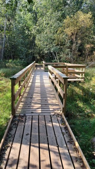 Bridge with handrails. In the middle of the bridge there is a widened section on the right and a bench at the edge of the widened section.