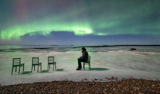 A man sits on a chair with three empty chairs in front of him, the northern lights blazing in the background.