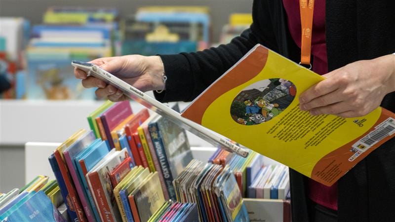En person håller i en barnbok i ett bibliotek eller en bokhandel. Personen har en svart tröja och ett orange halsband. I bakgrunden och förgrunden syns färgglada böcker på hyllor. På bokens baksida finns tecknade figurer och text.