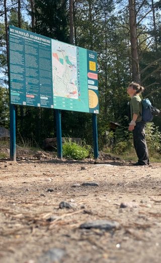 : Opasteen edusta on tasattua maapohjaa. Opaste on tumman vihreä ja siinä on tekstiä sekä kartta. Oikeassa alakulmassa on qr-koodi, jonka kautta opasteen voi kuunnella