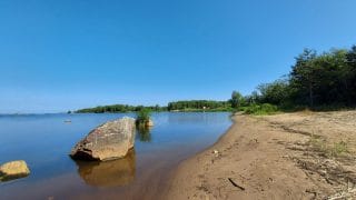 Hiekkarantaa ja tyyni vedenpinta. Vedessä rantaviivan lähellä on kiviä. Keskellä oikeassa reunassa näkyy metsää. Taivas on sininen-