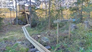 Lintulava on rakennettu siirtolohkareen päälle. Lavalle vie kahden lankun levyinen pitkospuu ja portaat. Pitkosten oikealla puolella on puustoa.