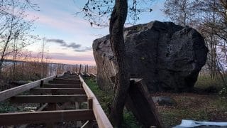 Oikeassa reunassa siirtolohkare, jonka vasemmalta puolelta kulkee keskeneräiset pitkospuiden rakenteet. Ilta-aurinko on maalannut taivaan osin vaalenapunaiseksi.