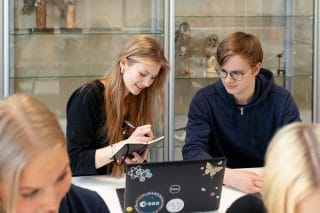 Kaksi nuorta opiskelee. Tyttö kirjoittaa muistikirjaan ja poika katsoo.